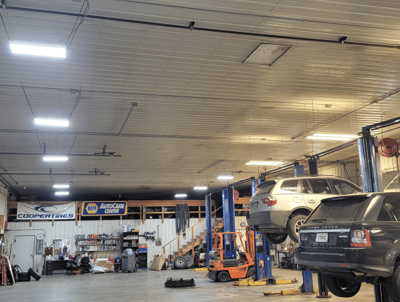 Scheduled Maintenance at Sioux Empire Automotive Service Center in Tea, SD. A spacious auto repair shop with two cars lifted on hydraulic lifts. The shop has various tools, equipment, and shelves stocked with supplies. Bright overhead lights illuminate the area, and several banners, including Cooper Tires and NAPA AutoCare Center, are visible.