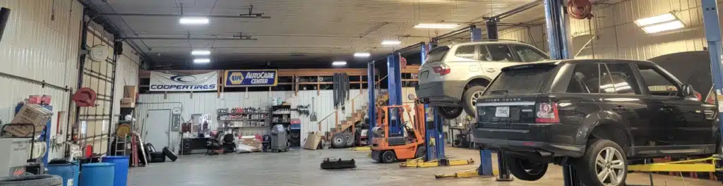 Auto repair shop interior with two cars raised on hydraulic lifts. Various tools and equipment are visible around the spacious area, along with storage shelves, a desk, and stairs leading to a loft area. Hanging banners display Cooper Tires and Napa AutoCare logos.