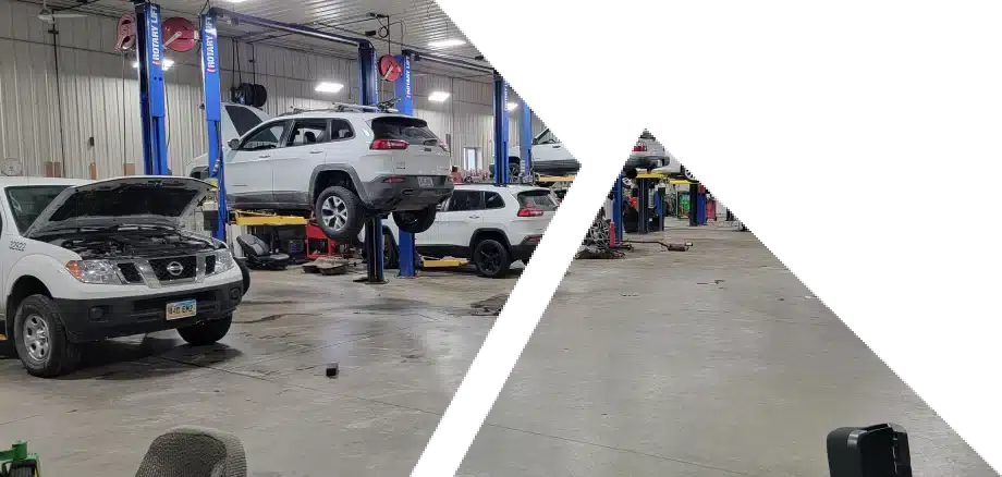 A spacious car repair garage with several vehicles being serviced. Two cars are elevated on hydraulic lifts with one having its hood open. Tools and equipment are scattered around the workshop, and several more vehicles are visible in the background.