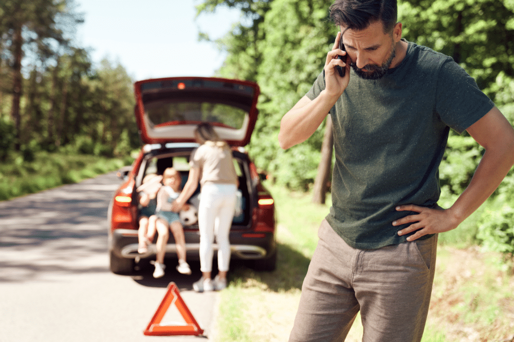 Tires, auto repair in Tea, SD at Sioux Empire Automotive Service Center. Man on phone looking concerned next to a broken-down car with hazard triangle placed on the road, while a woman and two children sit by the open trunk.