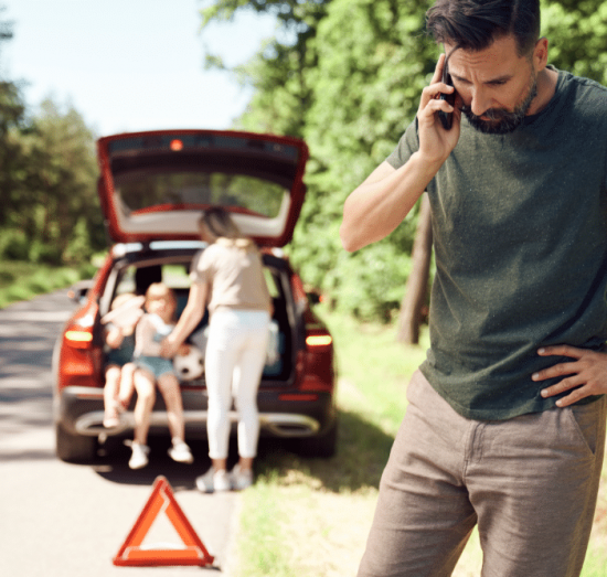 Tires, auto repair in Tea, SD at Sioux Empire Automotive Service Center. Man on phone looking concerned next to a broken-down car with hazard triangle placed on the road, while a woman and two children sit by the open trunk.